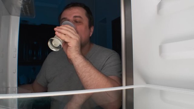 adult man opens refrigerator at night, takes glass of kefir and drinks it.