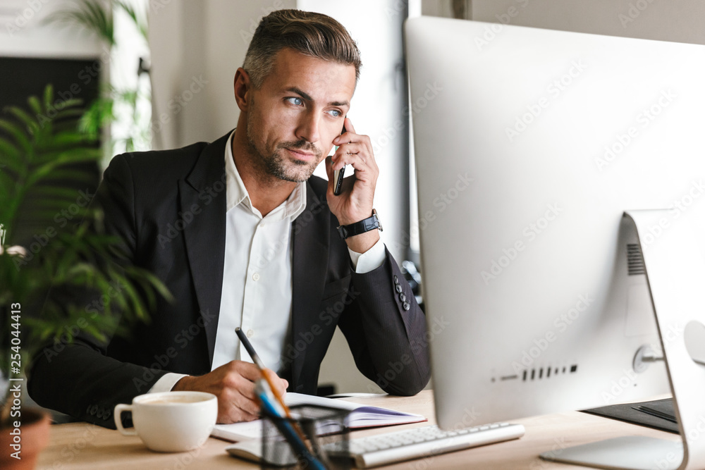 Poster Image of serious businessman talking on cell phone while working on computer in office