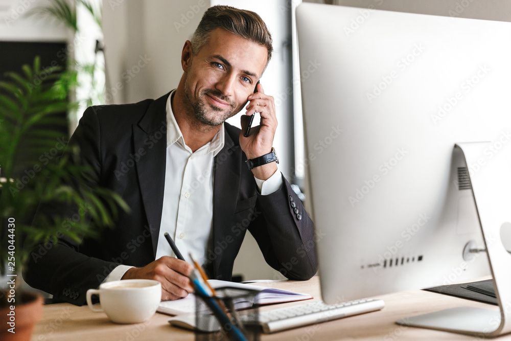 Poster image of happy businessman talking on cell phone while working on computer in office