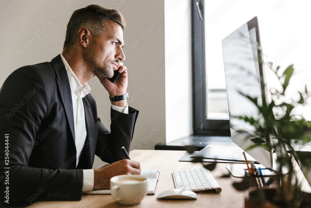 Poster image of successful businessman talking on cell phone while working on computer in office