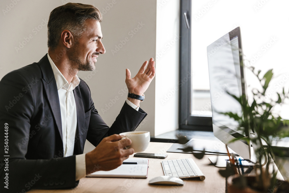 Poster image of joyful businessman drinking coffee while working on computer in office
