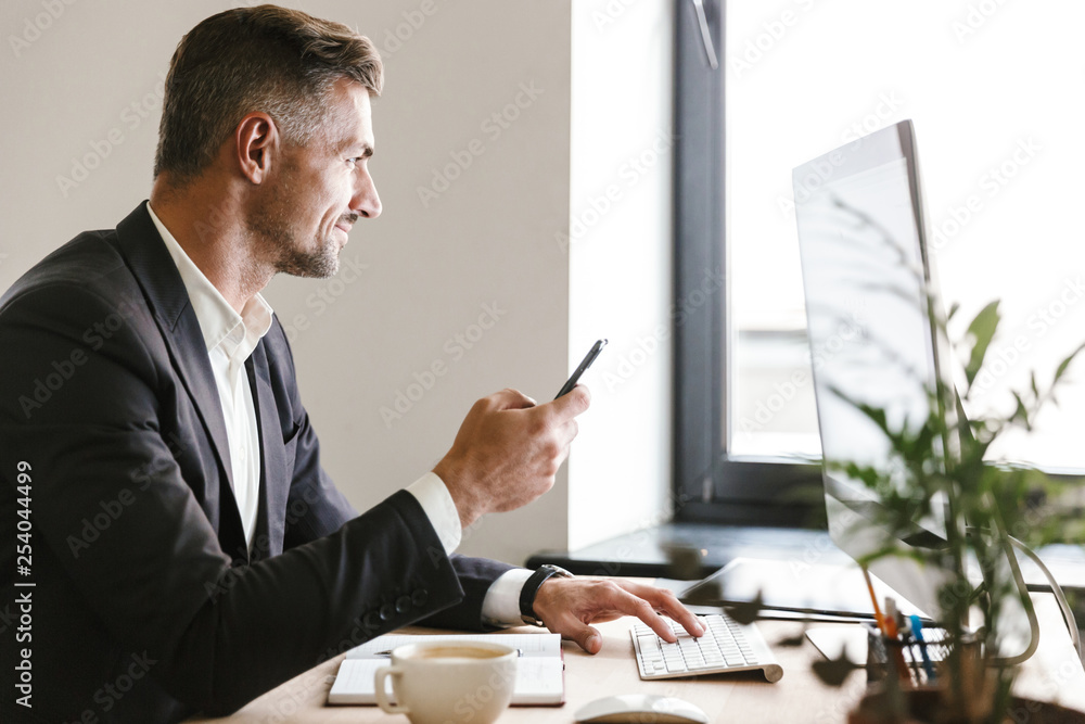 Poster portrait of successful businessman using mobile phone while working on computer in office