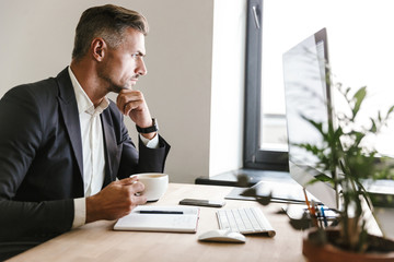 Image of concentrated businessman drinking coffee while working on computer in office