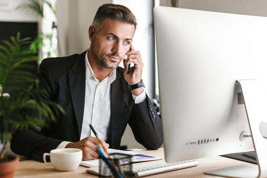 Image of serious businessman talking on cell phone while working on computer in office