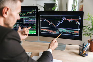 Photo of serious businessman sitting at table in office and working with graphics and charts on computer