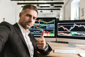Photo of confident man taking selfie while working in office on computer with graphics and charts at screen