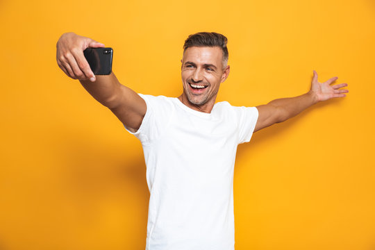 Image of good-looking man 30s in white t-shirt laughing and taking selfie photo on mobile phone