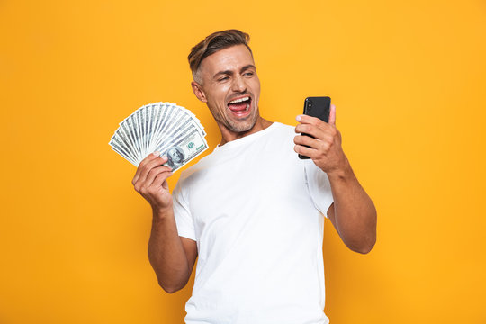Image of brunette guy 30s in white t-shirt holding cell phone and bunch of money