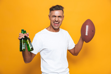 Image of joyful man 30s in white t-shirt drinking beer and holding rugby ball while standing isolated