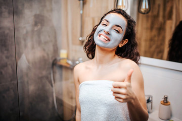 Young sporty dark-haired beautiful woman doing morning evening routine at mirror. Cheerful happy model look on camera and msile. She has face mask. Woman hold big thumb up.