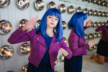 Fashion freak. Portrait of glamour synthetic girl, fake doll with empty look and blue hair is sitting in the studio. Stylish beautiful woman in purple jacket near light bulbs. Fashion and beauty