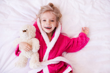 Fototapeta premium Happy kid girl 3-4 year old holding teddy bear lying in bed wearing bathrobe closeup. Looking at camera. Good morning. Top view