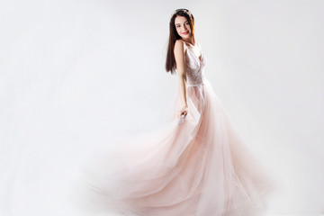 young brunette sexy girl in a white wedding boudoir dress and jewellery in hairstyle is smiling and dancing like swan princess on a white wall background , spring, bride concept