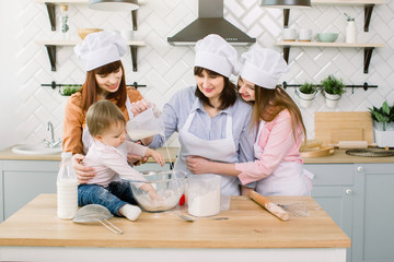 Cute Little Girl and Her Beautiful Mom, Aunt and Grandmother in Aprons and hats having fun While pouring milk to flour and Kneading the Dough in Modern Kitchen at Sweet Home. Women baking at kitchen