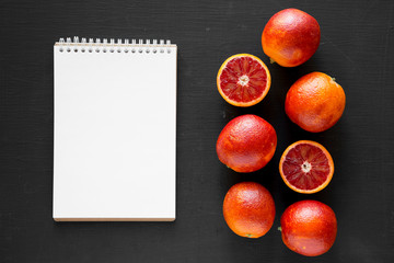 Whole and halved blood oranges, blank notepad on black surface, top view. Flat lay, overhead, from...