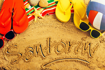 Summer concept of sandy beach, colorful thongs shoes, sunglasses, ball and inscription santorini