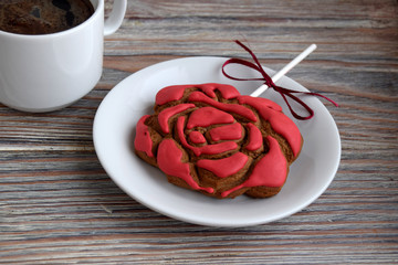Gingerbread in the form of a flower of a red rose is on a white plate. The cup with coffee is on a wooden table