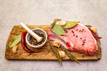 Raw pork steak with spices and dried herbs on vintage wooden board. Salt, garlic, hot pepper, rosemary, bay leaf with ceramic mortar and pestle on a stone background, close up.