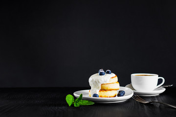 cottage cheese pancakes one on another topped with cream, blueberries and sugar powder and cup of coffee, fork and green mint leaves on black background, Horizontal with copy space for text or design
