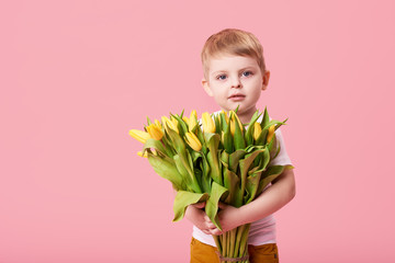 Adorable smiling child with spring flower bouquet looking at camera isolated on pink. Little toddler boy holding yellow tulips as gift for mom. Copy space for text 