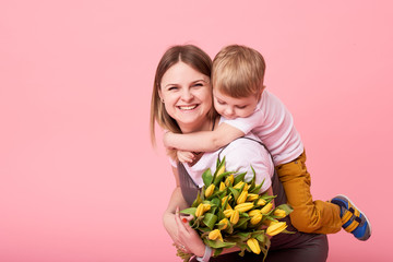 Young mother hugs his little son sitting on the floor against a pink background. Mom holds a...