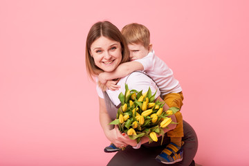 Young mother hugs his little son sitting on the floor against a pink background. Mom holds a bouquet of spring yellow flowers. Care and relationships and family concept. Mothers day