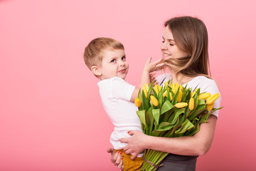 Fototapeta na wymiar Young mother hugs his little son sitting on the floor against a pink background. Mom holds a bouquet of spring yellow flowers. Care and relationships and family concept. Mothers day