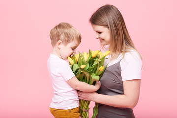 Fototapeta na wymiar Young mother hugs his little son sitting on the floor against a pink background. Mom holds a bouquet of spring yellow flowers. Care and relationships and family concept. Mothers day