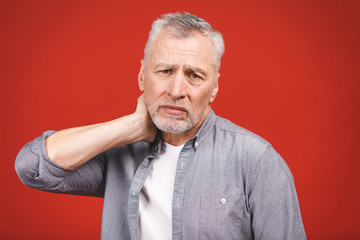 I need a massage. Frustrated senior man in casual clothing holding hand on his neck while standing against red background.