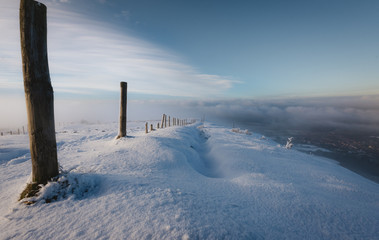 A line of Wooden Posts down a snowy hill into the valley below covered in fog