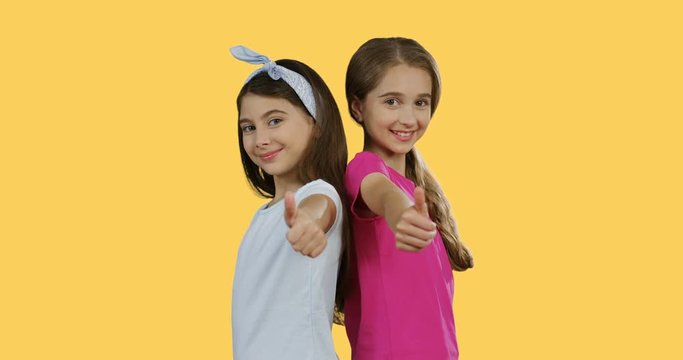 Portrait of the pretty teenagers girls giving her thumb up and smiling while standing back to back on the yellow wall background.