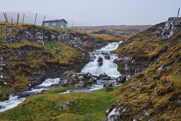 Wild creek or stream with waterfall cascade between fences of sheep pasture or grazing land with herdsman house during the cold, foggy, místy, rainy day in spring on Faroe islands in atlantic ocean.