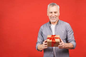 Mature senior man showing a gift with red ribbon as a present isolated against red background.