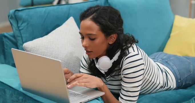Close up of the young mulatto attractive woman typing on the laptop computer and then smiling to the camera while lying on the couch in the living room.