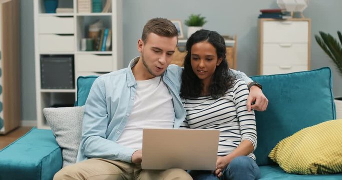 Mixed-races young happy couple sitting on the sofa in the living room and watching something on the laptop computer. Boyfriend hugging girlfriend.