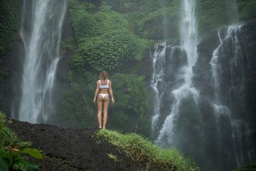 Fototapeta na wymiar back view of slim female in white bikini standing under great tropical fall,