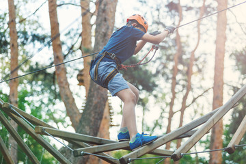 Sporty, young, cute boy in white t shirt spends his time in adventure rope park in helmet and safe equipment in the park in the summer. Active lifestyle concept