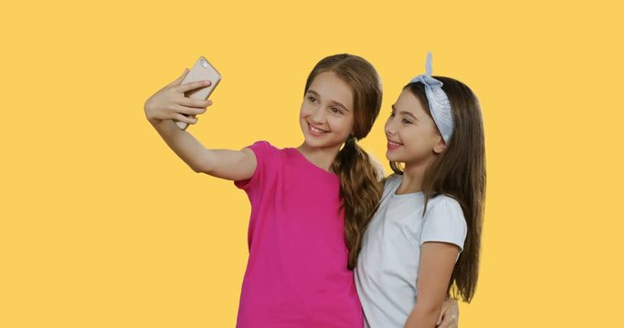 Caucasian friendly teens girls making selfies on the smartphone and posing while standing on the yellow background.