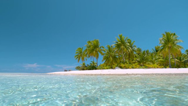 SLOW MOTION, LOW ANGLE, COPY SPACE: Glassy ocean water rolls past the picturesque tropical sandy beach and lush bright green palm trees. Cinematic view of paradise island nature under the clear sky.
