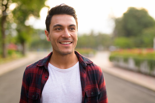 Face of happy young Hispanic hipster man thinking at the park