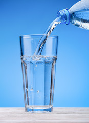 Sparkling water pours from  bottle into  large glass. Blue background, white table. Copy space.