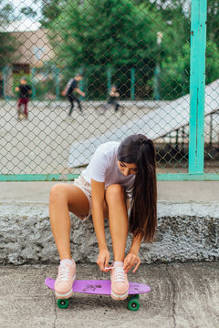Summer lifestyle image of trendy pretty young girl sitting next to the skateboard coart with her plastic skateboard and tying shoelaces.