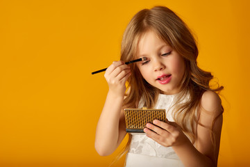 delight, caucasian, attractive, little girl, applying eyeshadow, using a brush, making, doing professional makeup on a yellow background