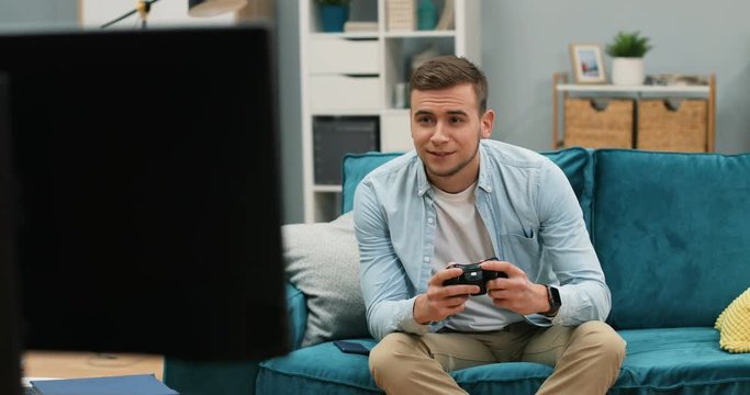 Caucasian young cheerful man sitting at home on the couch and playing videogame with a joystick in front of the TV screen.
