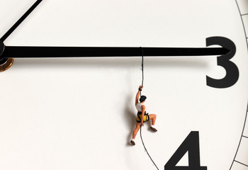 A miniature climber hanging by a rope on a black watch needle. - obrazy, fototapety, plakaty