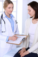 Fototapeta premium Young woman doctor and patient at medical examination at hospital office. Blue color blouse of therapist looks good. Medicine and healthcare concept