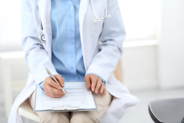Female doctor writing up medication history records form on clipboard, while sitting at the chair. Physician at work in hospital or clinic. Healthcare, insurance and medicine concept