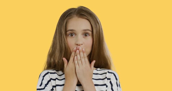 Close up of the blonde teenager girl with long hair in the striped blouse closing her mouth with hands and doing oops grimacing face. Yellow background.