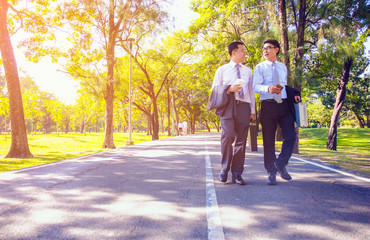 Fototapeta premium Businessman, They are walking on road in park. They are talking business. He is drinking coffee and His friend holding business bag. Photo concept business and relax time.
