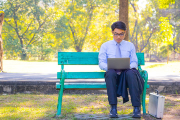 Fototapeta premium Businessman, He is sitting on bench in park. He is play notebook and thinking about business. He is search internet and relax time. Photo concept business and relax time.